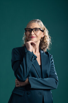 Senior woman wearing teal blazer black glasses, gold-hoops, holding chin, gazing upward in studio