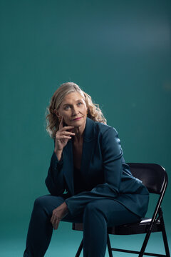 Mature woman sitting on black metal folding chair in studio, wearing tailored teal suit, pondering