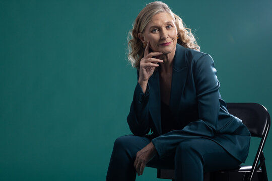 Mature woman sitting on black folding chair in portrait studio, wearing teal satin suit, copy space