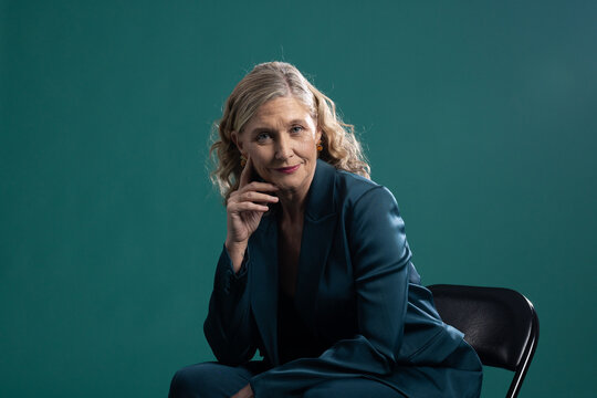 Senior woman sitting on folding chair touching chin in studio wearing teal suit, amber studs