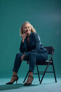 Senior woman sitting on black folding chair in studio wearing teal pantsuit and ankle-strap heels