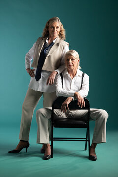 Senior female partners posing in studio on teal backdrop with metal folding chair, wearing suits
