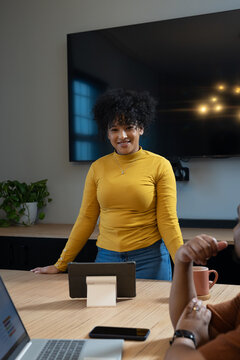 African American team leading in office, woman leaning on table, mustard top, jeans, tablet, laptop