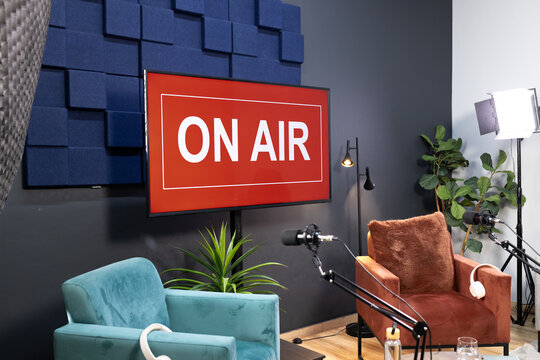 Wall-mounted monitor showing red ON AIR sign while boom microphones and chairs arranged in studio