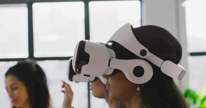 Diverse female team raising fingers after cue, tapping white VR headsets during demo near window