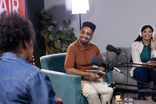 Diverse hosts and guest sitting in studio, man wearing orange polo holding notebook beside mics