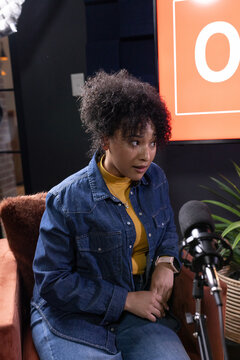 Woman sitting in studio armchair wearing denim jacket and mustard top, speaking into microphone