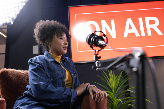 African American woman in denim jacket sitting recording studio speaking into mic with ON AIR sign