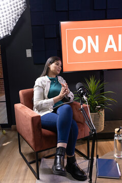 African woman sitting in rust armchair in studio with ON-AIR monitor, in jacket, speaking into mic