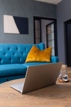 Silver laptop sitting open on wooden coffee table, glass of water, teal sofa with yellow pillow