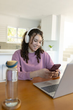 Teenage female sitting in kitchen in purple sweatshirt with headphones, scrolling phone near laptop