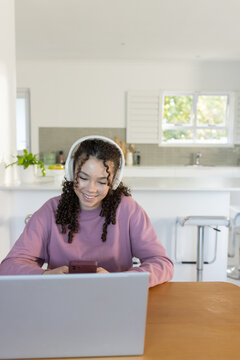 African American teen sitting at kitchen table, mauve sweatshirt, headset, using phone and laptop