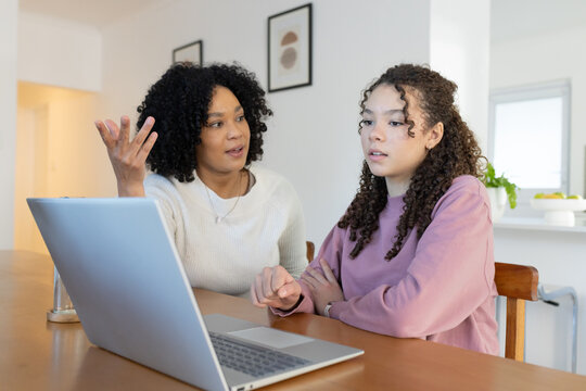 African American mother and teen daughter in knits sitting at wood dining table using silver laptop