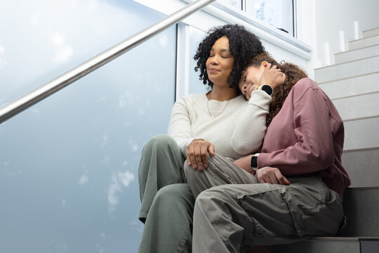 African American mother and teenage daughter sitting on stairs cradling head with smartwatch