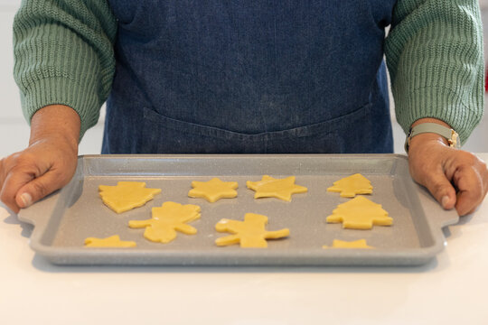 Senior African American woman wearing denim apron holding tray with cookie cutouts on white counter