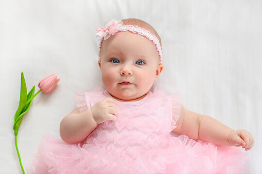 Baby girl with blue eyes wearing a pink ruffled dress and headband lies on a white blanket beside a pink tulip flower