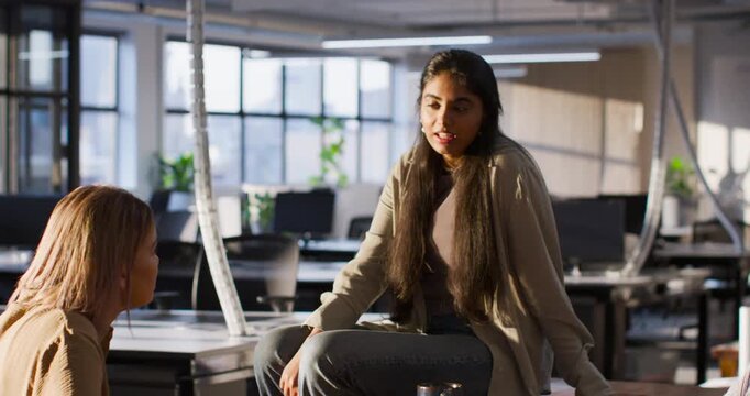Two female coworkers talking, speaker in cardigan replying after listening and gesturing at desk