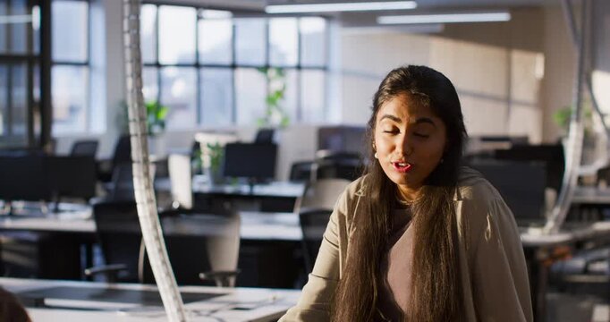 Indian woman sitting at desk in open office wearing khaki jacket reacting to offscreen peer joking