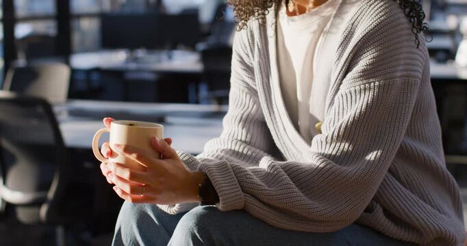 African American woman cupping mug after off-frame question, rolling sleeves and speaking in office