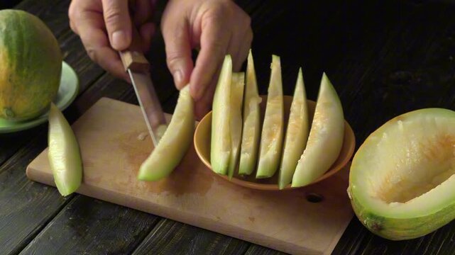 Hands slicing fresh melon into even wedges on a wooden cutting board, with a whole melon and a plate in the background, showcasing the preparation of a healthy snack in a kitchen setting
