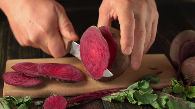 Hands of a person slicing a fresh beetroot on a wooden cutting board, with vibrant red slices and green leaves visible, showcasing the preparation of healthy ingredients in a kitchen setting