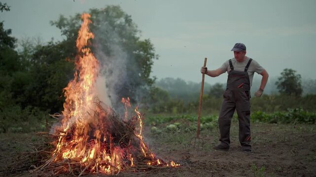 Farmer in gray overalls tending to a large fire made of branches in a rural field, observing the flames and smoke rising against a backdrop of trees and cloudy sky during early evening