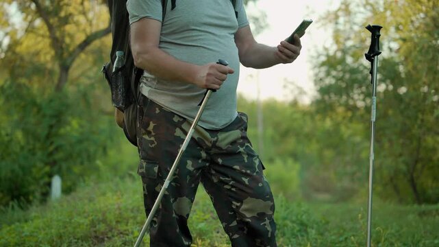 Hiker in camouflage pants and gray shirt stands on a hiking trail, checking his smartphone while holding trekking poles, surrounded by lush greenery and trees in a natural outdoor environment