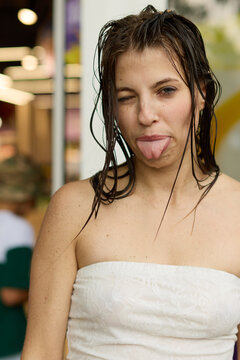 Portrait of a silly young woman with wet hair sticking out her tongue and winking, dressed in a white strapless lace top while celebrating a fun summer outdoor water festival.