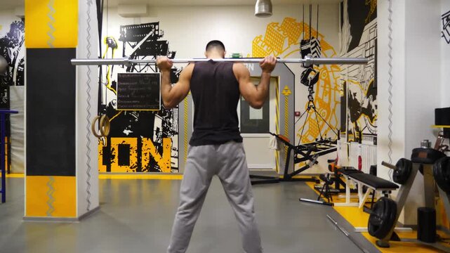 Muscular athlete sits down with a barbell at modern gym. Young strong sportsman doing weightlifting exercises at sport club. Athletic guy working out at fitness centre. Healthy active lifestyle