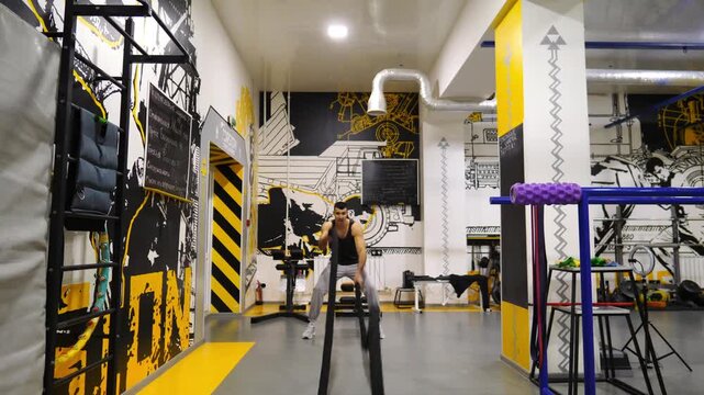 Young muscular sportsman training with battle ropes at modern gym. Strong sporty man exercising in fitness club. Athletic guy doing hard and intense workout. Concept of sport and active lifestyle