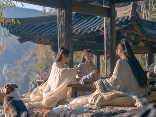 Fototapeta premium Three people in flowing ancient hanfu share tea beneath a weathered wooden pavilion, mountains and a small dog in the background.