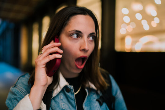 Shocked Caucasian female student listening on smartphone with open mouth expression outdoors, receiving unexpected mobile banking alert or cybersecurity notification in city at night