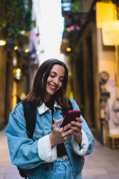 Cheerful Caucasian female freelancer looking down at smartphone screen in European alley, managing remote freelancing client messages digital workflow and project schedule outdoors