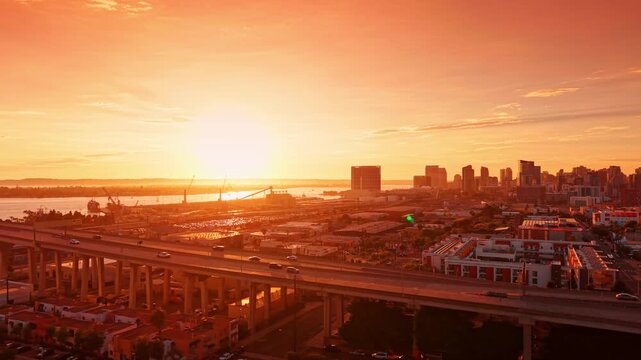 Warm sunlight dazzling the scenery of the city. Cars move by the Coronado Bridge at sunset. Port cranes in the backdrop. California, USA.