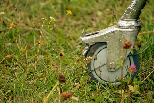 A metallic swivel caster wheel with a brake mechanism stands on vibrant green lawn surrounded by wild flowers outdoors during daytime.