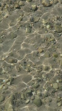 Klares Wasser im Hochformat: Stille Wellenbewegung in Slow Motion mit Lichtspiegelung gibt den Blick frei auf braunen Sand und Steine am Seegrund