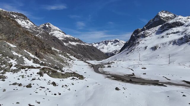 mountain pass road winds through open valley cars travel past roadside buildings power line poles julier pass snowbound slopes graubunden switzerland high altitude alpine crossing route