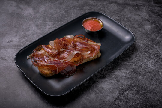 Traditional Spanish toasted bread topped with cured serrano ham served alongside crushed tomato sauce on a dark slate surface, Mediterranean cuisine