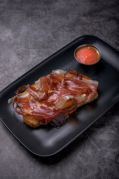Traditional Spanish toasted bread topped with cured serrano ham served alongside crushed tomato sauce on a dark slate surface, Mediterranean cuisine