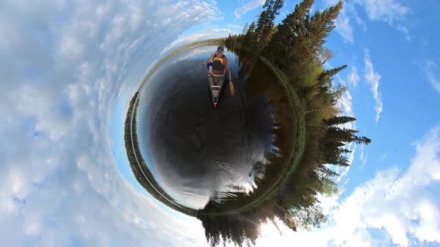 Wide angle globe like view of person paddling a canoe on a calm lake in a northern forest
