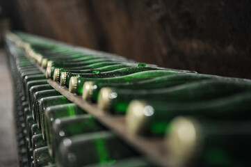 Rows of dusty green champagne wine bottles aging horizontally in a dark underground traditional stone cellar with shallow depth of field and warm dim lighting. © samy