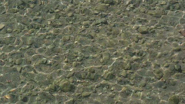 &Ouml;kosystem und Wassermenge im See: Glasklares, flaches Wasser in Slow Motion mit verzerrter Sicht auf Sand und Steinen am Grund. Sonnenlicht spiegelt sich in den Wellen einer klaren Wasseroberfl&auml;che 
