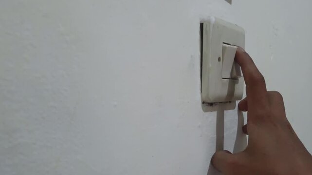 Hand pressing a wall-mounted light switch indoors, turning the electrical switch on and off for everyday household use.