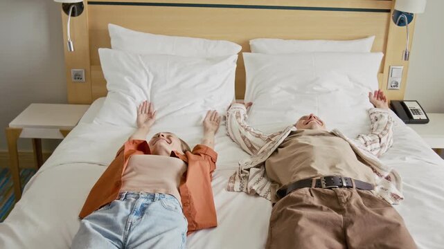 Caucasian young adult couple falling backwards onto hotel bed celebrating vacation arrival. Suitable for travel advertising, hotel booking campaigns, tourism promotion