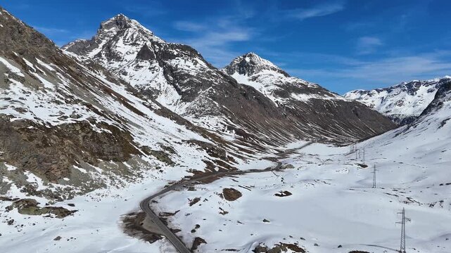 broad snowfield valley threads between towering peaks paved road winds along valley floor scattered pylons cross open terrain julier pass alpine crossing graubunden switzerland