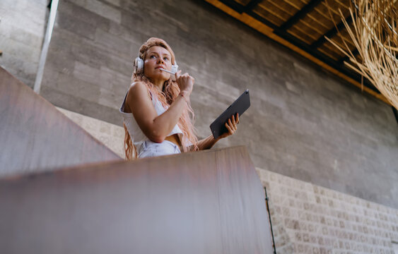Low angle view Caucasian female graphic designer in headphones holding stylus to chin thoughtfully, planning mobile app wireframe layout on touch tablet while standing on stairs