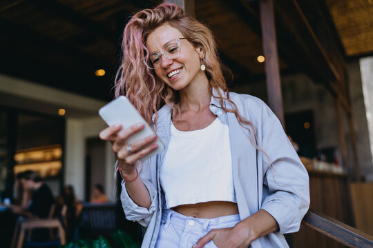 Caucasian female student in glasses smiling warmly while texting on smartphone, receiving positive online exam result notification standing in modern coworking studio workspace.