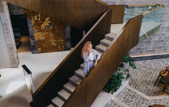 High angle view of Caucasian female IT specialist holding phone call conversation on staircase, discussing cloud software project details in modern open plan coworking studio venue