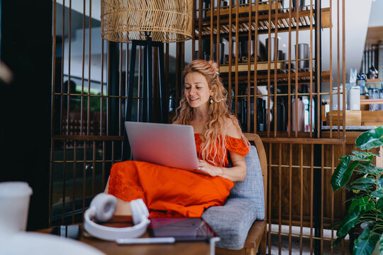 Caucasian female IT specialist in red dress sitting in armchair working on laptop computer, with wireless headphones and digital tablet on coffee table in modern open plan lounge workspace