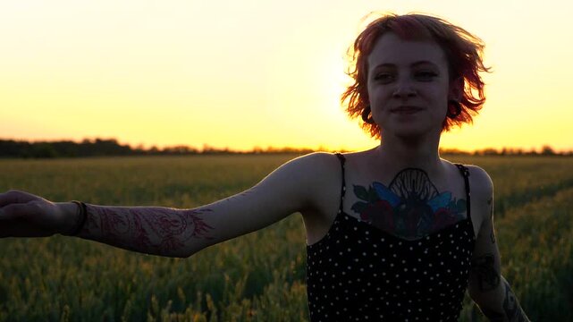 Happy female hipster with tattoos having fun during goes through green barley field at beautiful sunset. Cheerful punk girl in short black dress fooling around while walks among wheat meadow. Slow mo